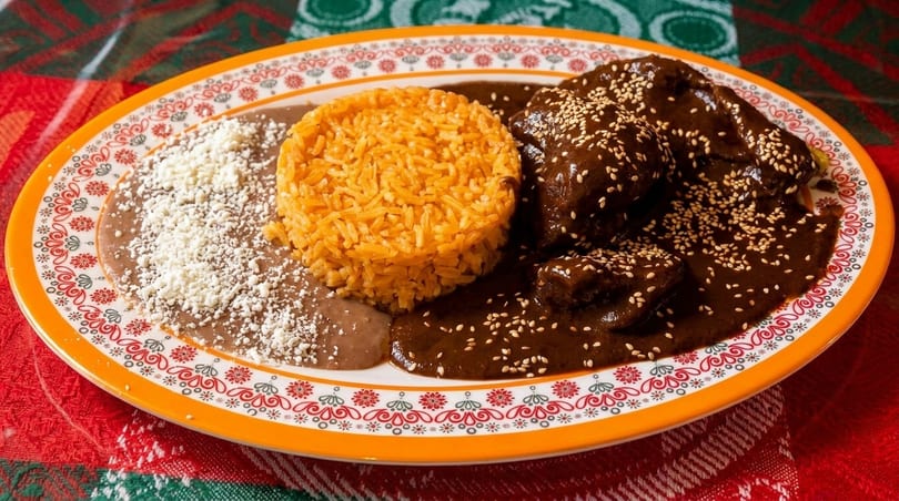 A plate of chicken mole with rice and refried beans, garnished with sesame seeds and cheese.