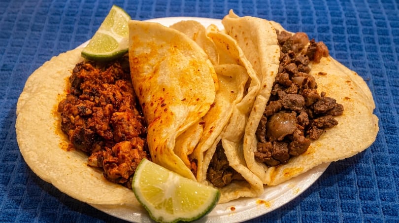 A plate of three tacos, two with dark meat and one with red spiced meat, garnished with lime wedges.