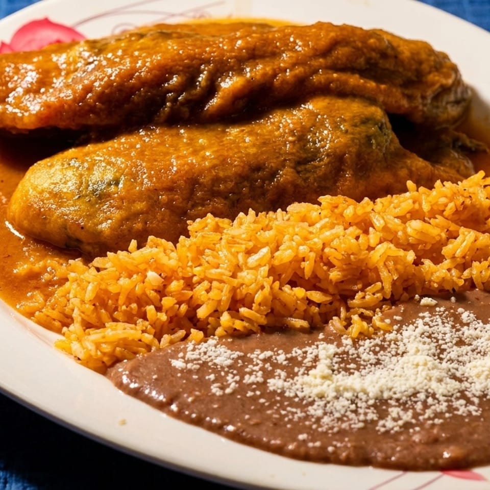 Two chile rellenos covered in red sauce, with a side of orange rice and refried beans.