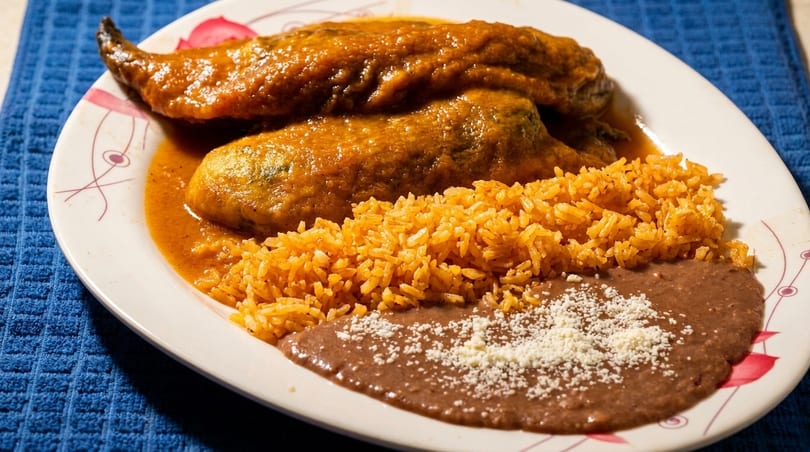Two chile rellenos covered in red sauce, with a side of orange rice and refried beans.