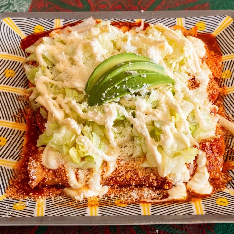 Enchiladas topped with shredded lettuce, cotija cheese, crema, and avocado slices on a patterned plate.
