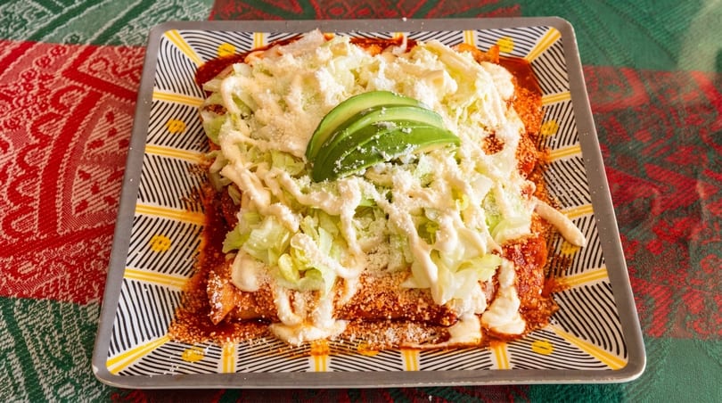 Enchiladas topped with shredded lettuce, cotija cheese, crema, and avocado slices on a patterned plate.