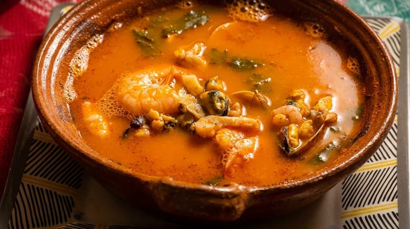 Close-up of a rustic bowl filled with vibrant orange seafood soup, featuring shrimp, mussels, and clams.