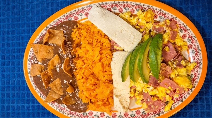 Mexican breakfast plate with refried beans, rice, scrambled eggs with ham, cheese, and avocado slices.